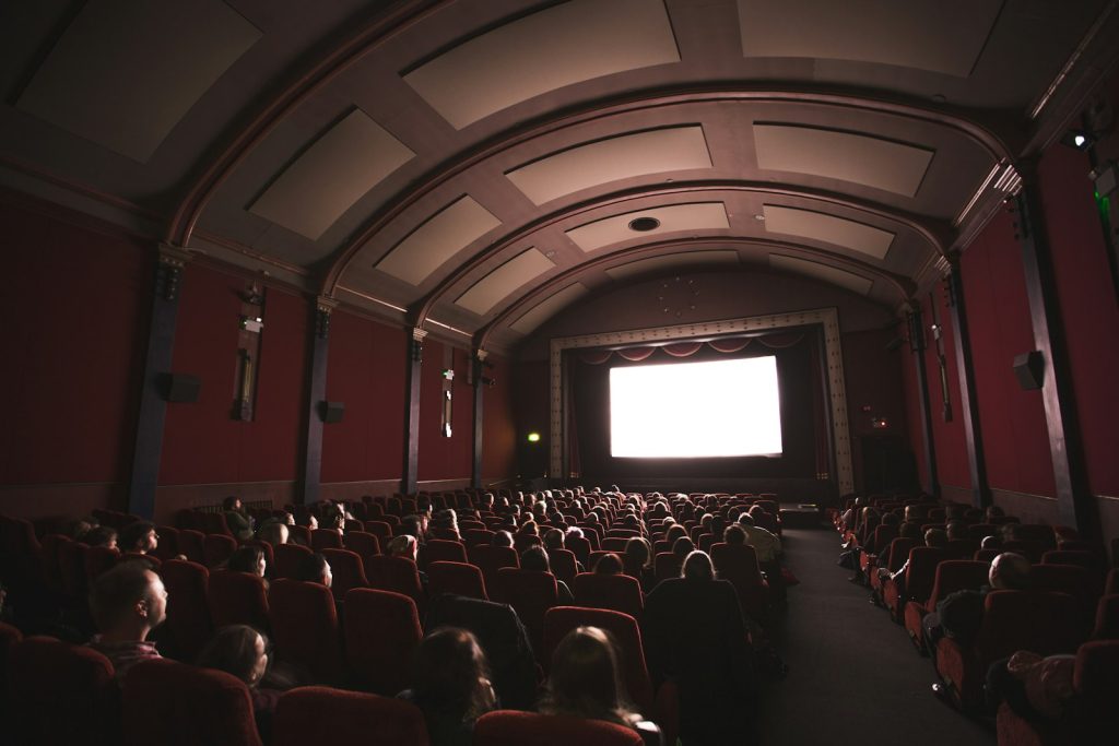 Audience in a dark cinema with red seats facing a bright screen